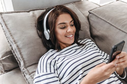 pretty African American woman laying on couch with headphones on using phone - recovery-related podcasts
