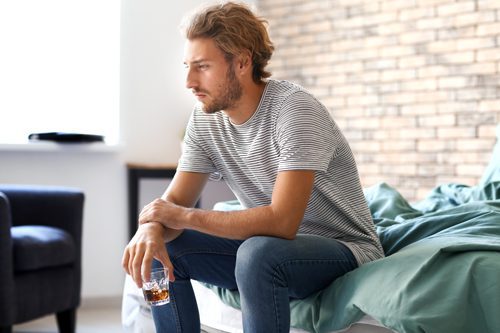 sad man sitting on bed at home holding a glass of liquor - joblessness and addiction