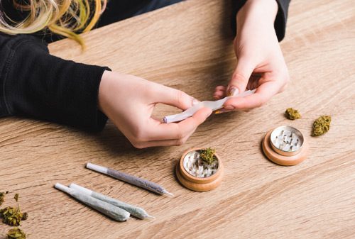 cropped shot of a woman with long blonde hair rolling joints of marijuana and using an herb grinder - marijuana