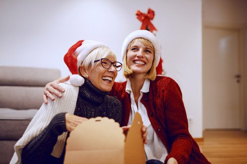 lovely senior woman and younger woman smiling wearing Santa hats and opening a gift together - express gratitude