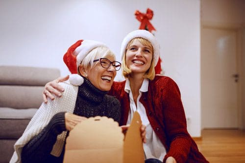 lovely senior woman and younger woman smiling wearing Santa hats and opening a gift together - express gratitude