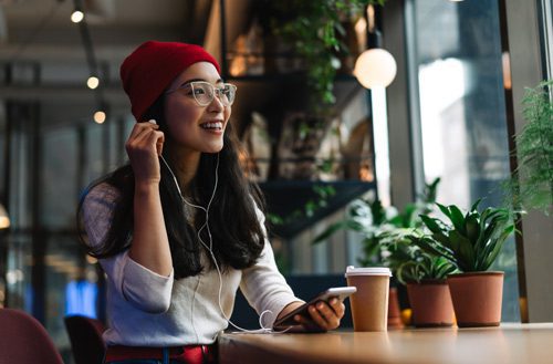 beautiful young Asian woman wearing glasses and a red knit cap, listening to headphones and smiling - music