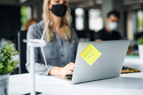 woman sitting at her work desk with a black cloth mask on; there is a sticky note on her laptop that says Welcome Back! - work after treatment