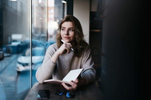 pretty young woman looking thoughtful and writing in a journal at a cafe near a large window that looks out onto the street - understanding stress