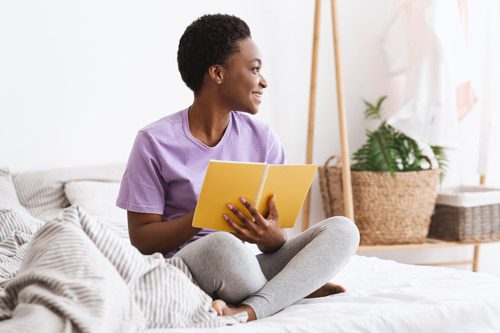 beautiful Black young woman sitting on her bed at home smiling, thinking about what to write in her journal - journaling prompts