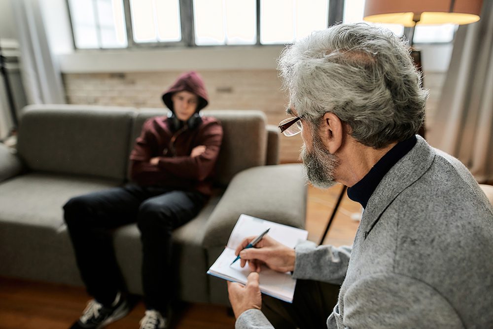 Inpatient addiction treatment Psychoanalyst taking notes in notepad during appointment with uneasy teenage boy sitting with crossed hands on couch in office. Psychotherapy concept