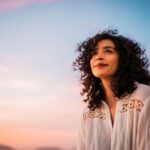 Woman with curly hair smiling gently while looking into the distance during sunset, with a pastel-colored sky in the background.