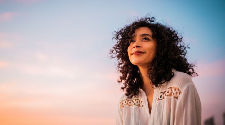 Woman with curly hair smiling gently while looking into the distance during sunset, with a pastel-colored sky in the background.