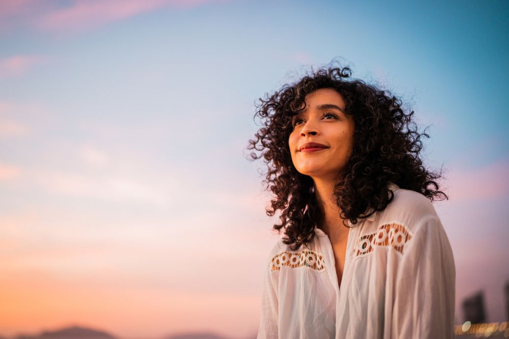Woman with curly hair smiling gently while looking into the distance during sunset, with a pastel-colored sky in the background.