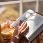 Person reading a book by a sunny window, with soft light casting shadows on the pages—conveying relaxation, reflection, and personal growth.