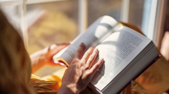 Person reading a book by a sunny window, with soft light casting shadows on the pages—conveying relaxation, reflection, and personal growth.