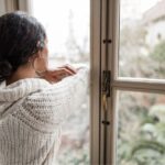 Woman in a cozy sweater standing indoors, resting her arms on a window frame and looking out thoughtfully at a blurred outdoor scene.