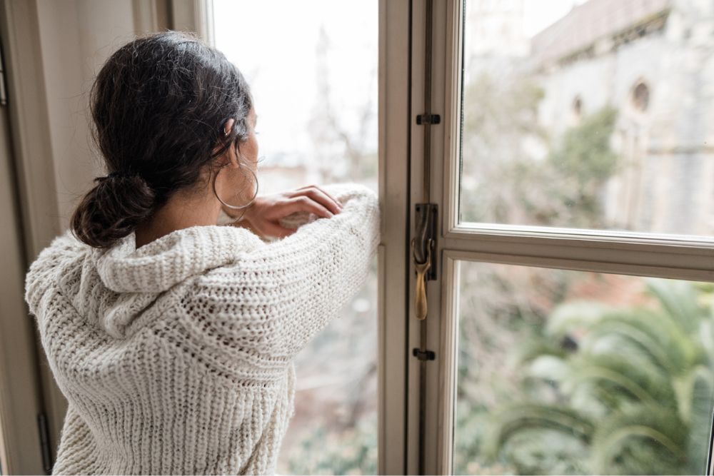Woman in a cozy sweater standing indoors, resting her arms on a window frame and looking out thoughtfully at a blurred outdoor scene.