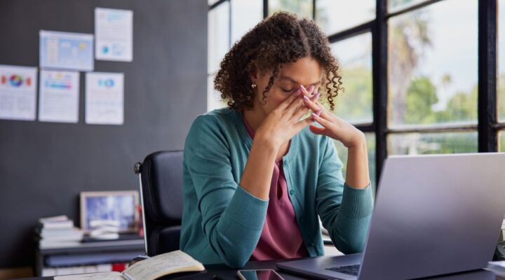Woman sitting at a desk with a laptop, holding her face in her hands in a gesture of stress or fatigue, surrounded by papers, a notebook, and a smartphone.