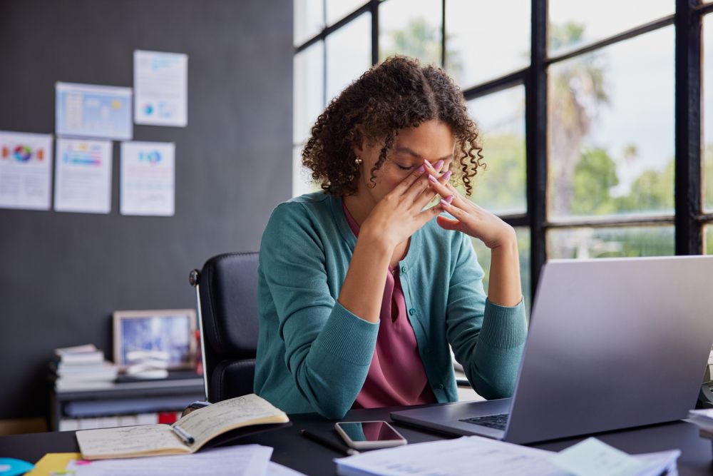 Woman sitting at a desk with a laptop, holding her face in her hands in a gesture of stress or fatigue, surrounded by papers, a notebook, and a smartphone.