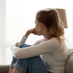 A young woman sitting on a couch with her knees drawn up, looking out the window thoughtfully, suggesting introspection, concern, or emotional struggle.