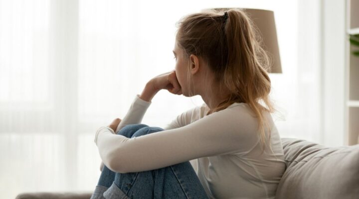 A young woman sitting on a couch with her knees drawn up, looking out the window thoughtfully, suggesting introspection, concern, or emotional struggle.