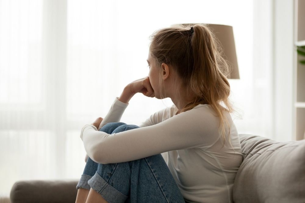 A young woman sitting on a couch with her knees drawn up, looking out the window thoughtfully, suggesting introspection, concern, or emotional struggle.