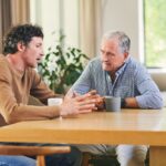 Middle-aged man speaking with an older man over coffee at a kitchen table, symbolizing support and mentorship in addiction recovery.