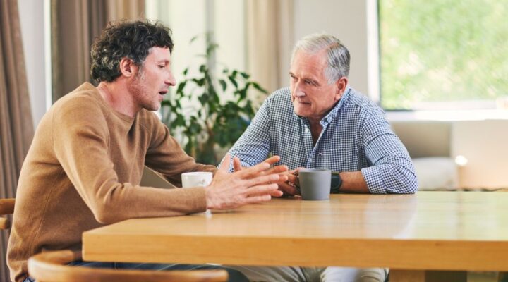 Middle-aged man speaking with an older man over coffee at a kitchen table, symbolizing support and mentorship in addiction recovery.