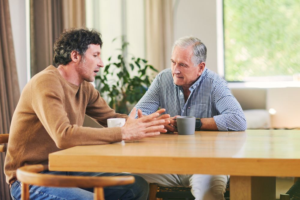 Middle-aged man speaking with an older man over coffee at a kitchen table, symbolizing support and mentorship in addiction recovery.