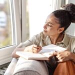 Woman sitting by a window, holding a journal and pen, looking outside thoughtfully while writing, suggesting reflection or mindfulness practice.