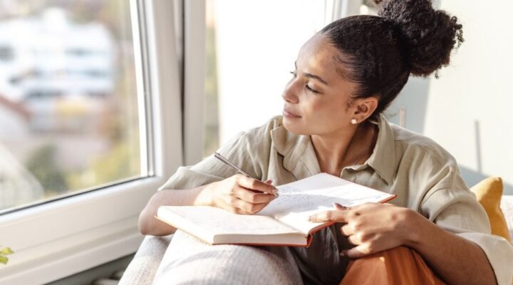 Woman sitting by a window, holding a journal and pen, looking outside thoughtfully while writing, suggesting reflection or mindfulness practice.