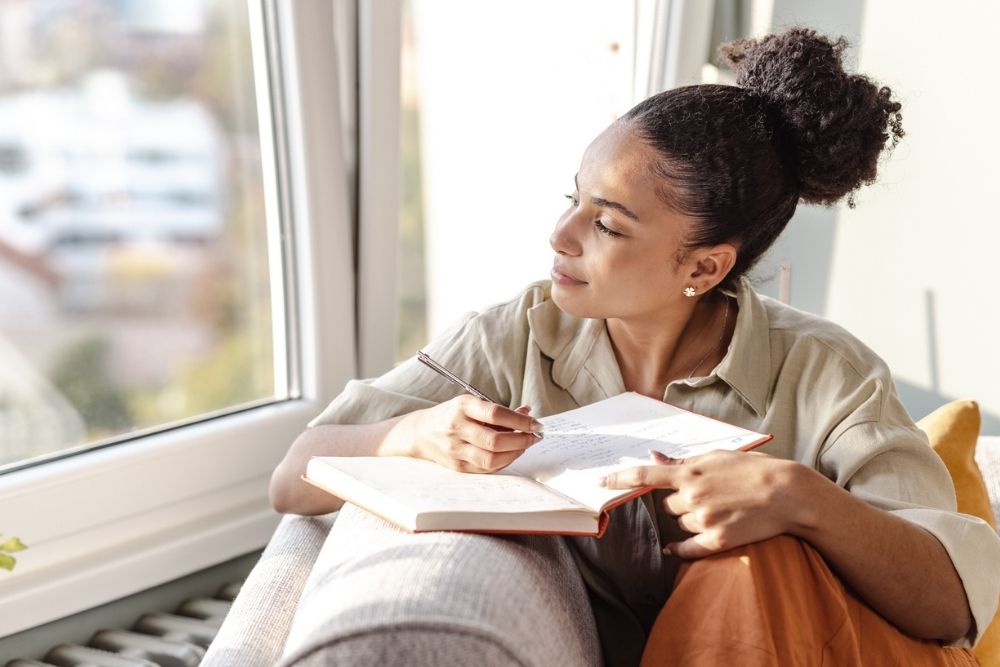 Woman sitting by a window, holding a journal and pen, looking outside thoughtfully while writing, suggesting reflection or mindfulness practice.