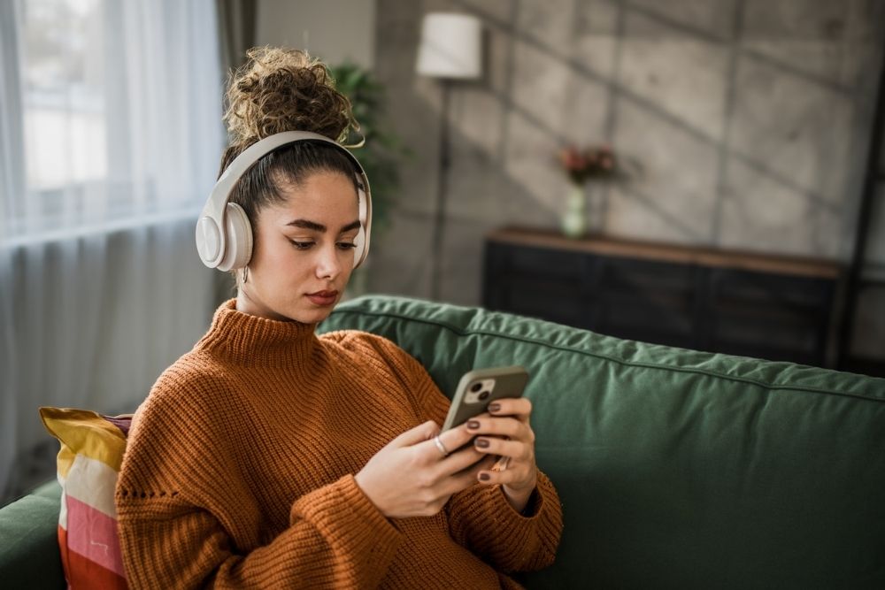 Excited young woman listening to music on wireless headphones over a mobile app while sitting on a comfortable sofa