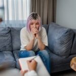 Young woman sitting on a couch with a thoughtful expression during a therapy session, while a counselor takes notes, symbolizing trauma therapy.