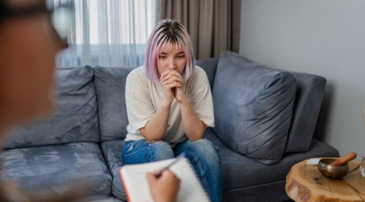 Young woman sitting on a couch with a thoughtful expression during a therapy session, while a counselor takes notes, symbolizing trauma therapy.