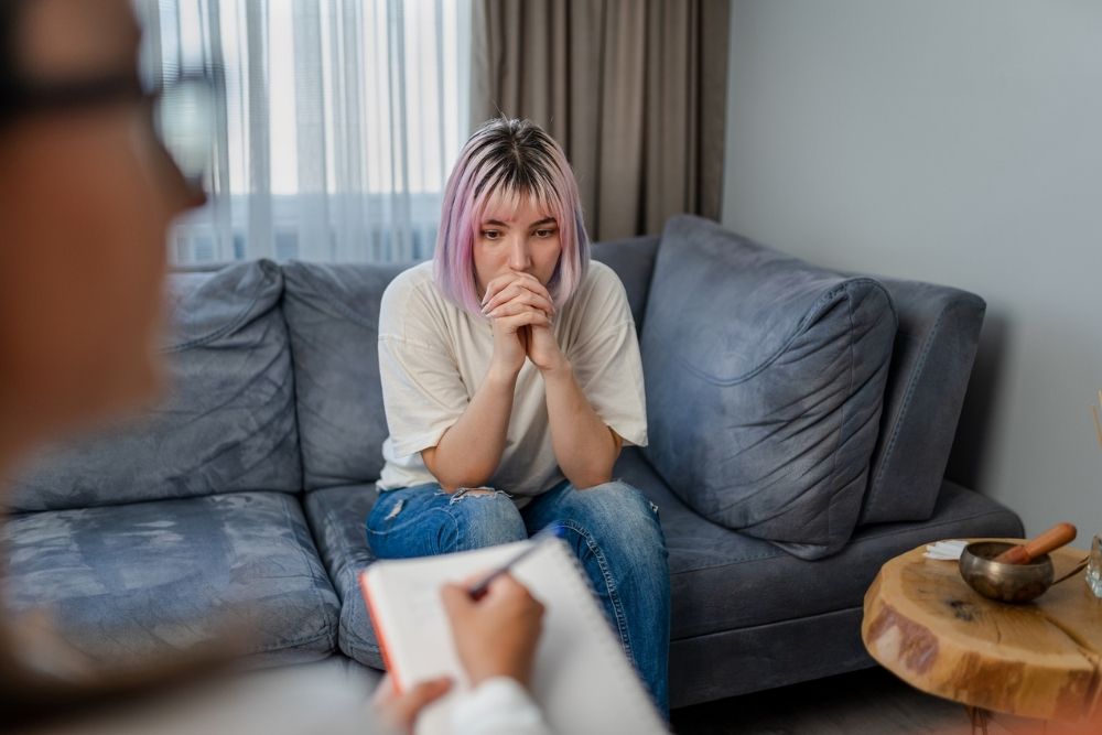 Young woman sitting on a couch with a thoughtful expression during a therapy session, while a counselor takes notes, symbolizing trauma therapy.