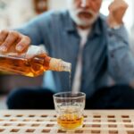 An older man sits on a couch pouring whiskey into a glass, symbolizing excessive drinking or alcohol use disorder.