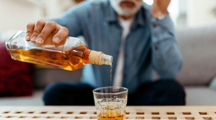 An older man sits on a couch pouring whiskey into a glass, symbolizing excessive drinking or alcohol use disorder.