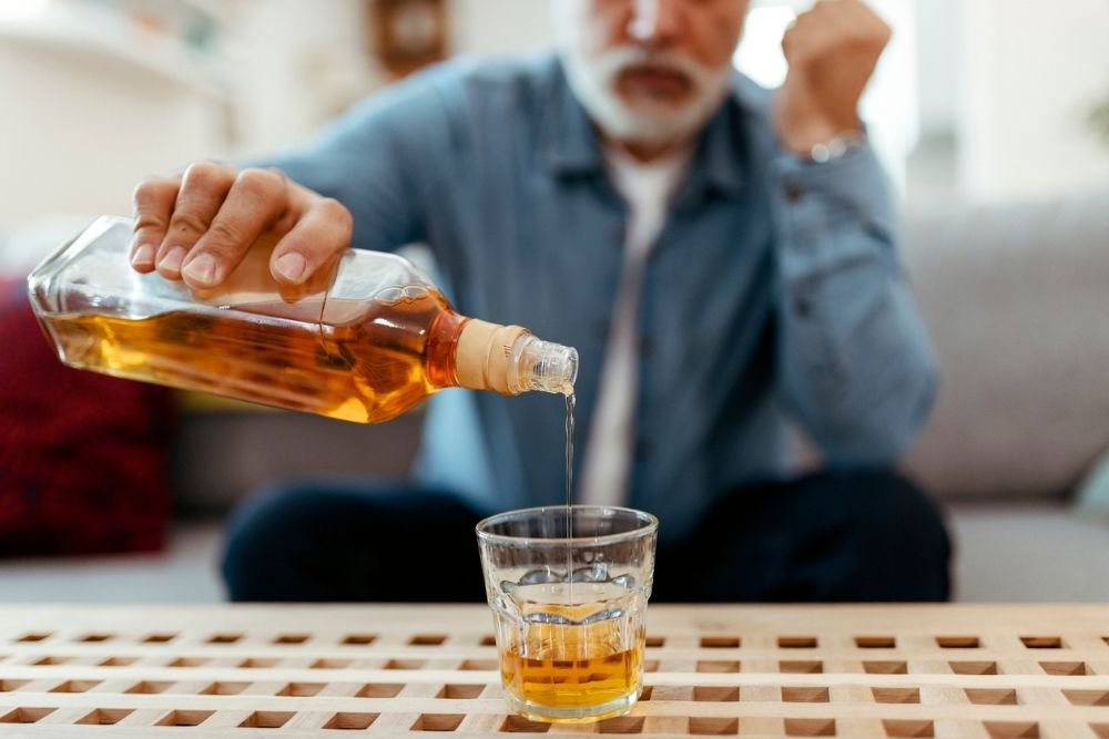 An older man sits on a couch pouring whiskey into a glass, symbolizing excessive drinking or alcohol use disorder.