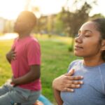 Two people practicing mindful breathing outdoors with eyes closed and hands on their chests, sitting on yoga mats in a peaceful park setting.