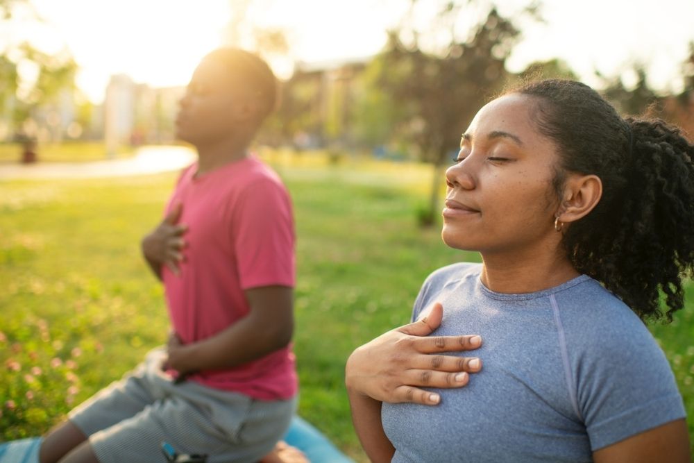 Two people practicing mindful breathing outdoors with eyes closed and hands on their chests, sitting on yoga mats in a peaceful park setting.