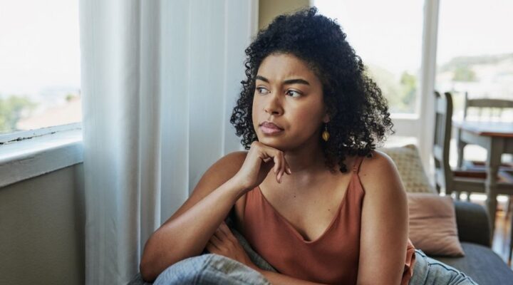 A woman sits indoors by a window, resting her chin on her hand and looking thoughtful, with a serious expression on her face.