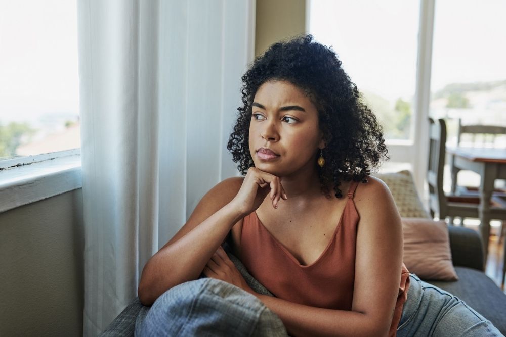 A woman sits indoors by a window, resting her chin on her hand and looking thoughtful, with a serious expression on her face.