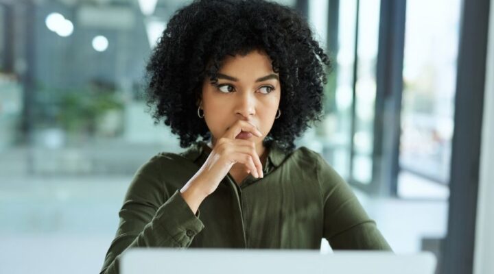 A woman sits at a desk with her hand near her mouth, looking thoughtful and concerned. She appears deep in worry or reflection in a bright, modern office setting.