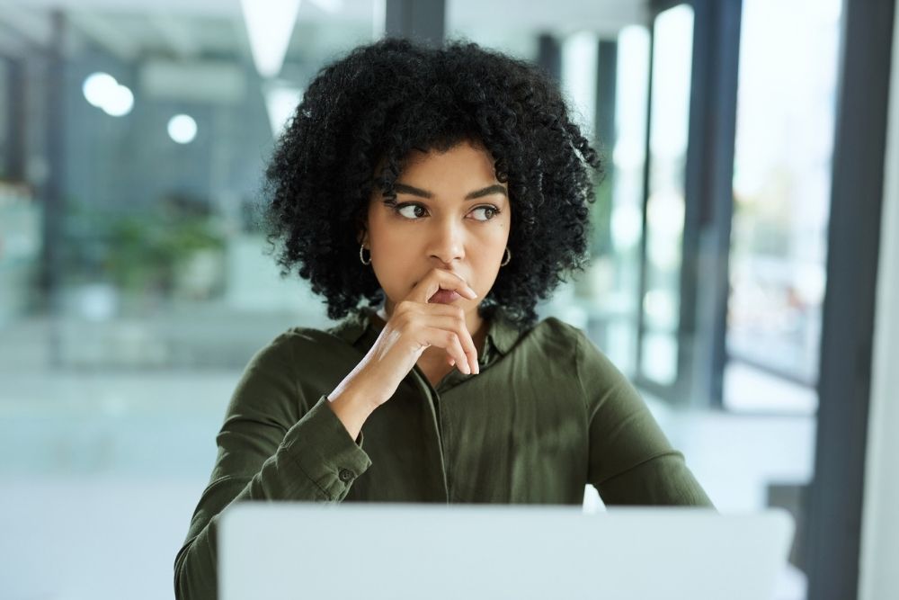 A woman sits at a desk with her hand near her mouth, looking thoughtful and concerned. She appears deep in worry or reflection in a bright, modern office setting.