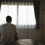 A man sits on the edge of a bed facing a window, shoulders slumped, conveying loneliness or depression in a quiet bedroom.