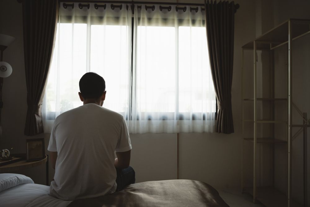A man sits on the edge of a bed facing a window, shoulders slumped, conveying loneliness or depression in a quiet bedroom.