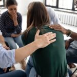 A support group sits in a circle. A person in a green sweater is comforted by others as they address grief during addiction recovery. The setting feels empathetic and caring.