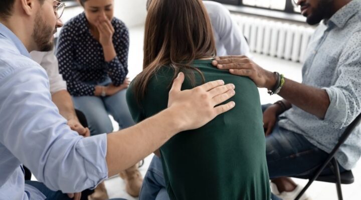 A support group sits in a circle. A person in a green sweater is comforted by others as they address grief during addiction recovery. The setting feels empathetic and caring.