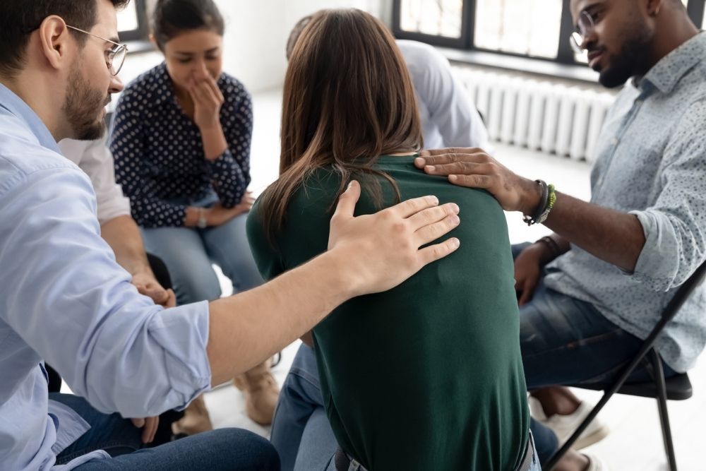 A support group sits in a circle. A person in a green sweater is comforted by others as they address grief during addiction recovery. The setting feels empathetic and caring.