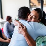 Two people hugging in a bright room with others in the background.