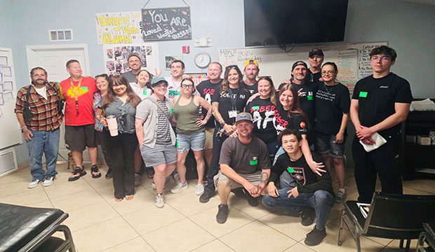 A cheerful group of about 20 people are gathered in a room with motivational posters on the walls. They are smiling, conveying a sense of camaraderie and positivity.