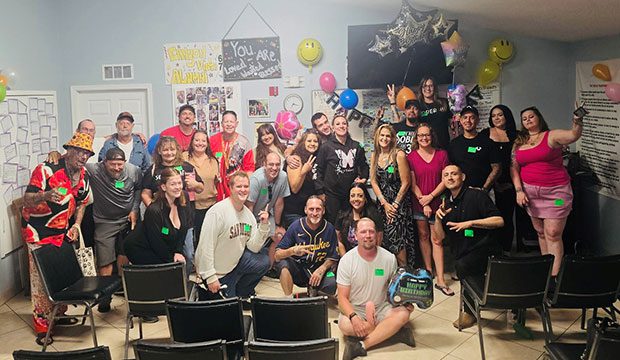 Group of people smiling at a celebration in a decorated room with balloons and posters. Casual setting, cheerful atmosphere, and chairs visible.